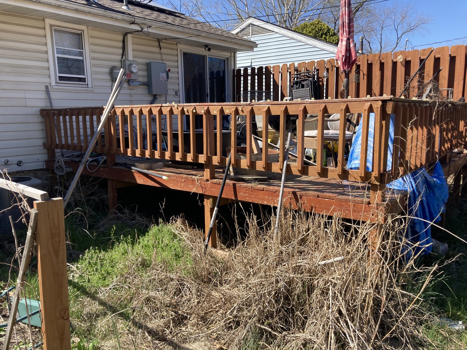 Existing deck seen from the yard, cluttered with furniture and a tarp, with dead brush grown up underneath