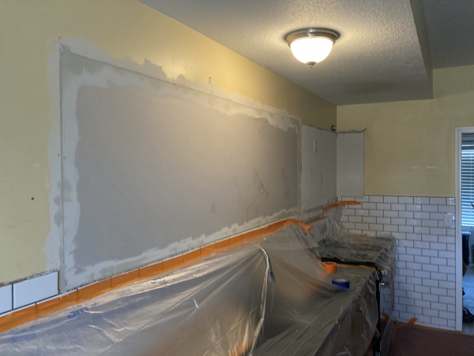 Kitchen wall with fresh drywall skim coat above subway tile, countertop covered in plastic.