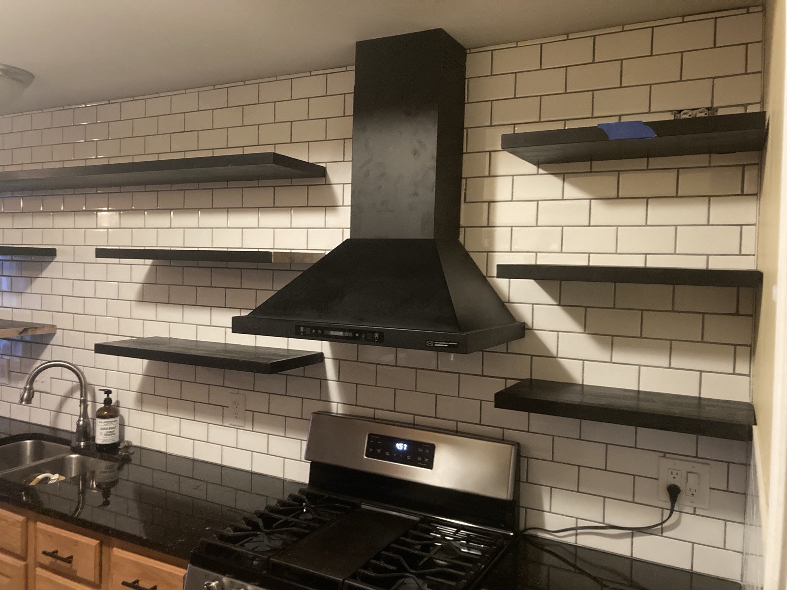 Finished dark wood floating shelves on either side of a black range hood over subway tile.