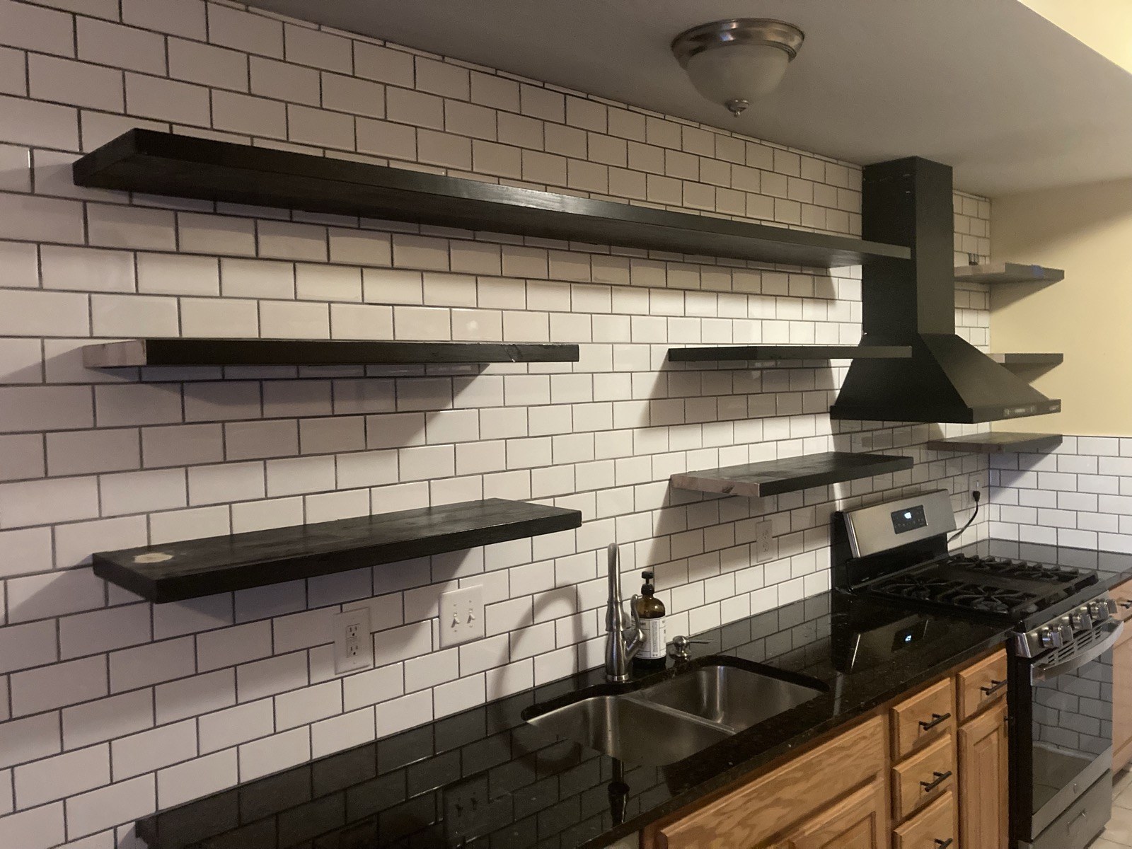 Finished kitchen with dark floating shelves and black range hood over white subway tile.