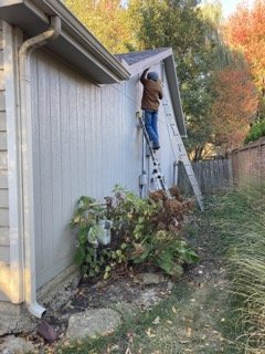 Person on ladder examining existing siding on gable end of house before repair