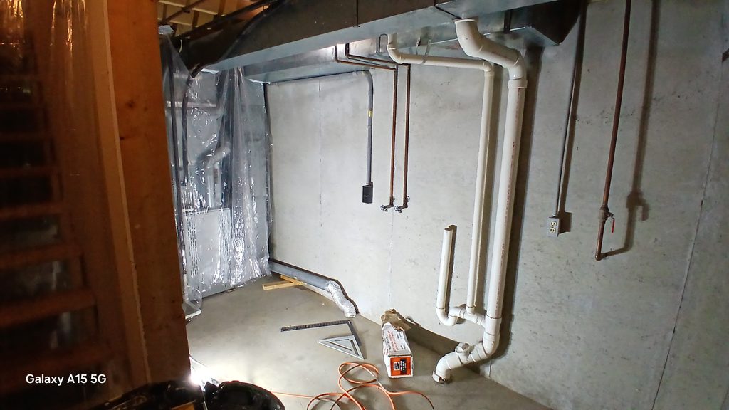 Unfinished basement corner showing plastic-wrapped water heater and exposed plumbing lines.
