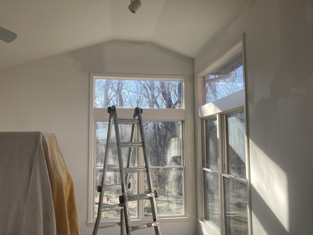 Sunroom corner with a tall arched window taped for paint and a ladder in place.