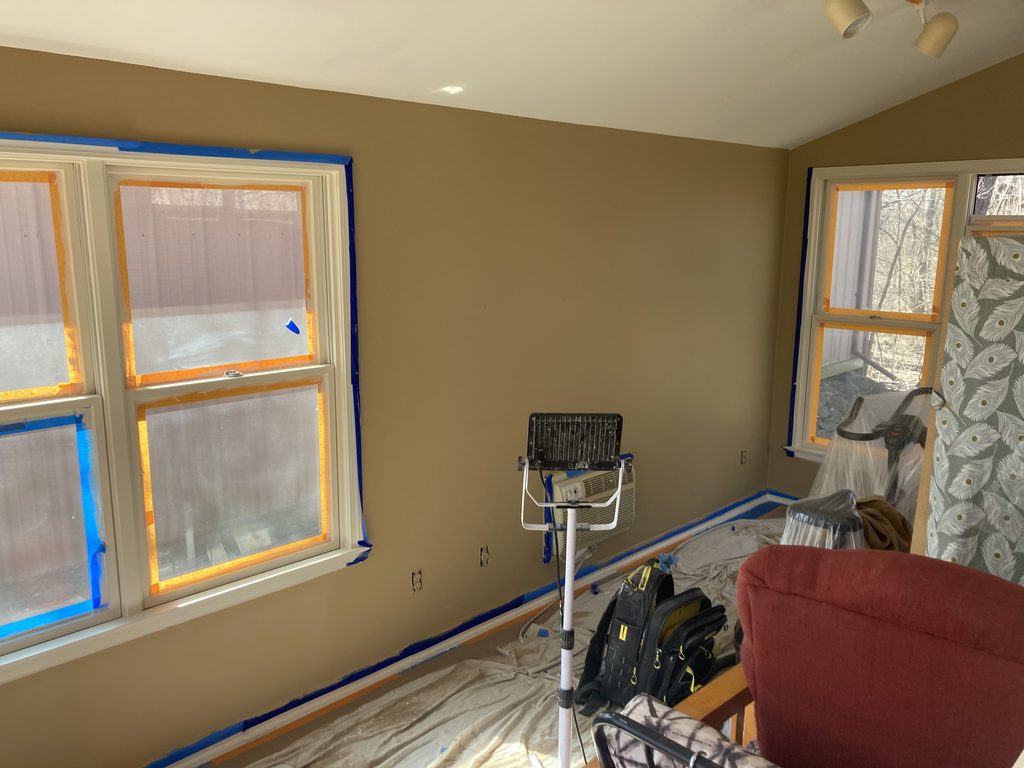 Sunroom wall freshly painted tan with windows taped and furniture covered nearby.