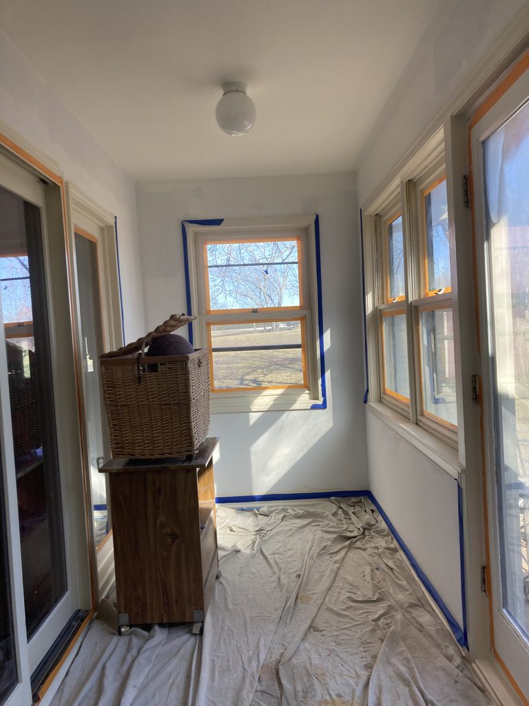 Sunroom with taped windows and a wicker basket staged on a wooden stand during the work.
