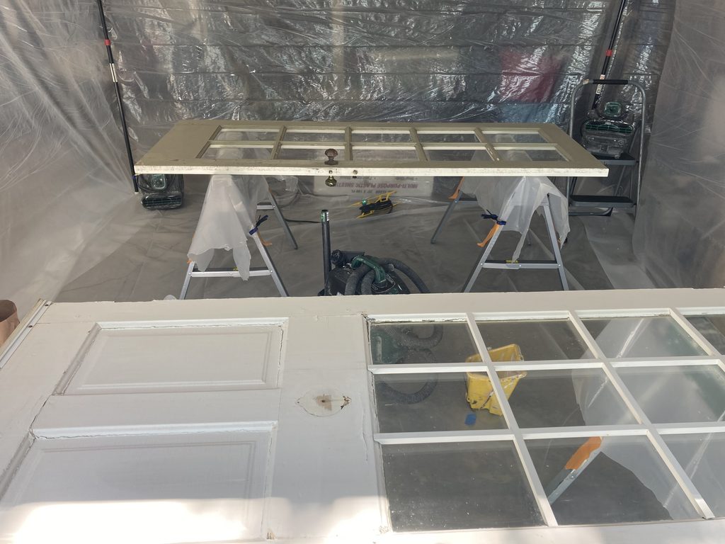 Second primed door on sawhorses inside a plastic paint tent, viewed from the opposite end.