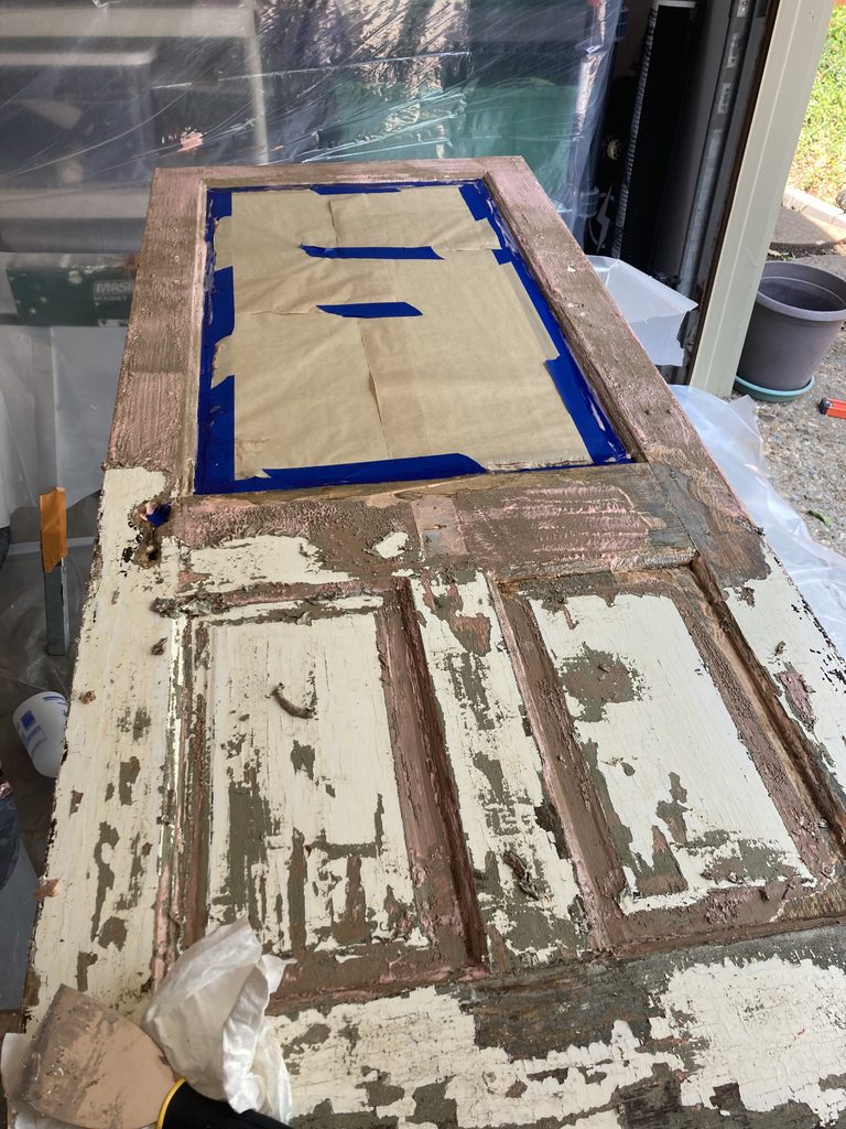 Multiple layers of peeling white and green paint on a door, with blue painter's tape marking a test area.