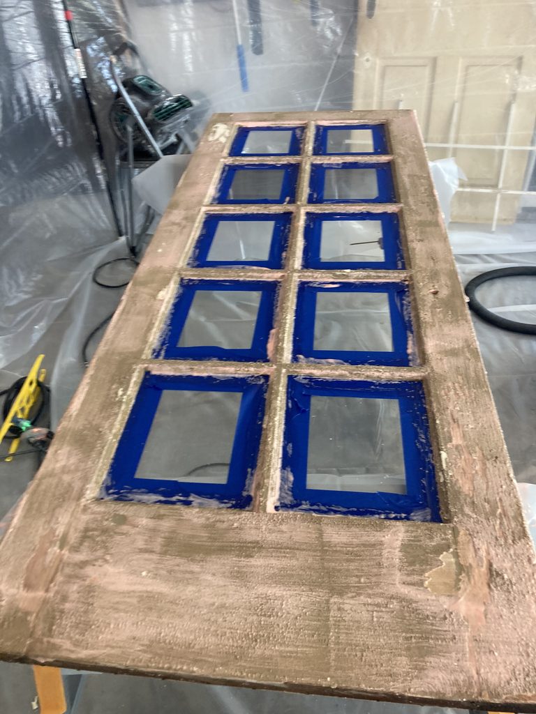 Ten glass window panes on a door masked with blue painter's tape, wood frame stripped to bare wood.