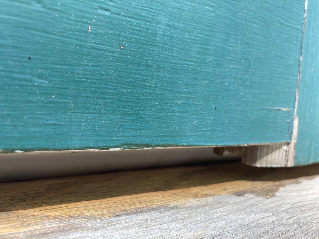 Close-up of teal paint and worn wood at the bottom corner of a door.
