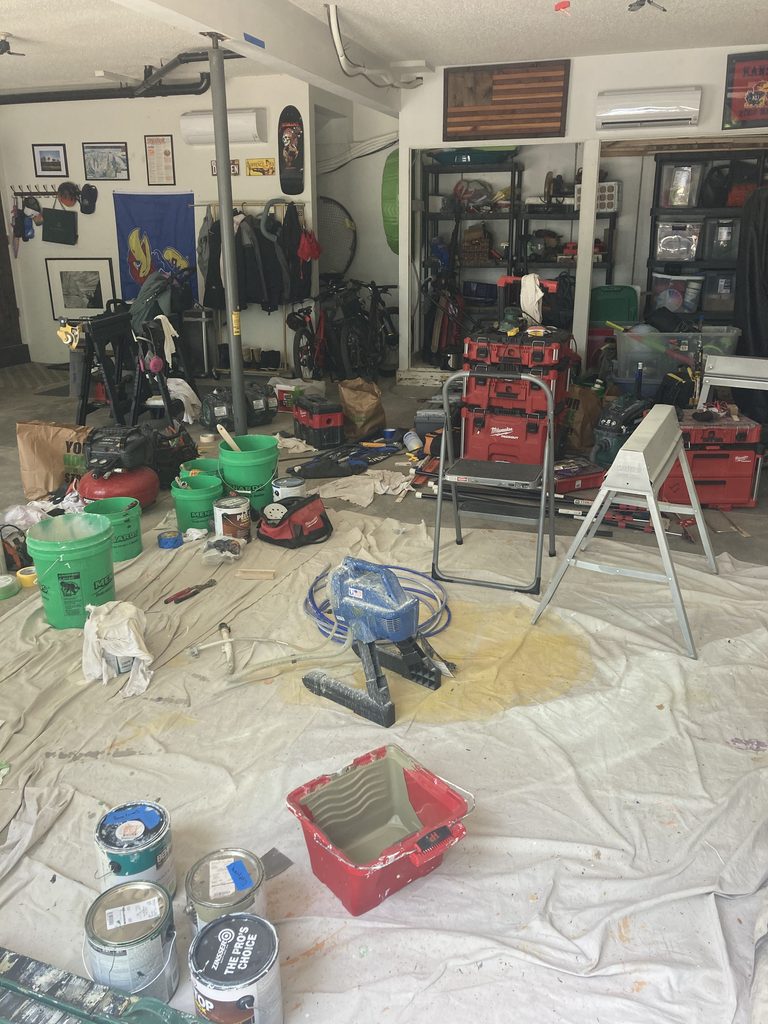 Garage floor covered with drop cloths, paint cans, tool boxes, and a paint sprayer.
