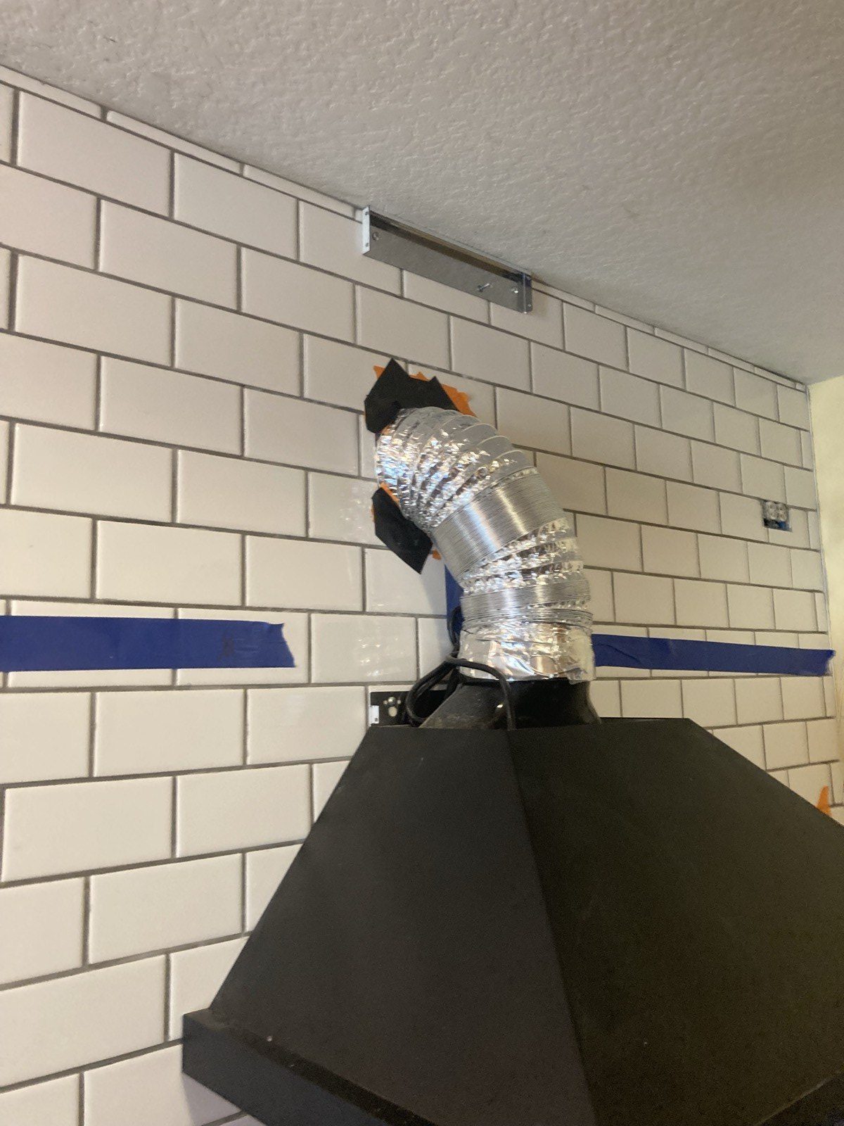 Close-up of a flexible metal duct running from the range hood through the tile wall.